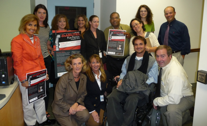 Pictured, a group of thirteen individuals of various genders, races, and ages pose for a picture, with three of them holding red, white, and black posters that announce the “disABILITIES Film Festival.”. 