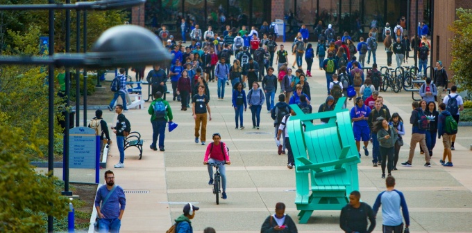 A birds’ eye view of Founders Plaza on UB’s North Campus. A blurry streetlight frames the left of the image with dozens of students moving in various directions around “Whippy,” a piece of public art made from bright aqua-colored plastic picnic tables. 