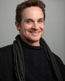 Director Michael Rembis, a white man with short, dark hair wearing a dark sweater and scarf against a gray portrait background, smiling at the camera. 