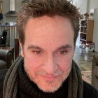 A color photo of the director, Michael Rembis. He is wearing a black turtleneck sweater and a grey scarf. He has short brown hair, hazel eyes, and brown scruff on his face. 