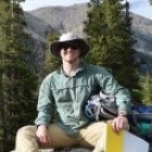 PhD student, Joe Tulenko sitting on a small boulder with trees and mountains behind him. 