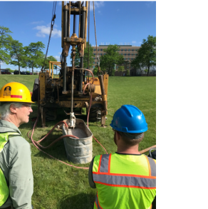 Two people wearing hardhats and green safety vests watch the large drill rig drill into the well.