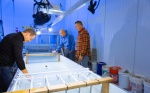 Zoom image: Left to right: Sean Bennett, geography, Adam Grodek, geography, and Kevin Cullinan, instructional support associate, gather around a flume that is used to research fish movement in February. This is installed in a research area of the Statler Commissary. Photographer: Douglas Levere