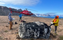 Geologists conduct field research on glacial deposits in Greenland, taking notes beside a large boulder with a red helicopter and Arctic mountains in the background.