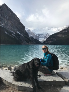 Richelle Allen-King and her dog Pearl sitting on a large rock in front of Lake Louise with a view of mountains in the distance. 