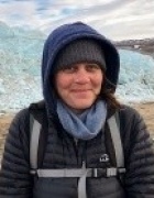Professor, Sophie Nowicki. Exploring glaciers and keeping warm. She is wearing a gray hooded coat and a light blue scarf. 