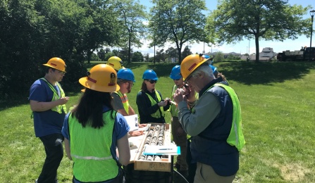 Field Camp Director, Professor Tracy Gregg talks with students around a table as they observe the first half of a 50 foot rock core. All are wearing hard hats and safety vests. 