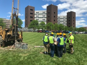 UB Geology Field Camp students getting ready to drill into the ground near Cooke Hall on North Campus. There are seven students all wearing hardhats and fluorescent green safety vests. 