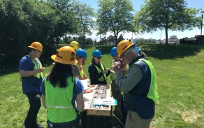 Field Camp Director, Dr. Tracy Gregg talks with students around a table as they observe the first half of a 50 foot rock core. All are wearing hard hats and safety vests. 