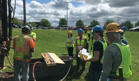 Students wearing hard hats and safety vest observe the first few meters of rock core coming out of the new well field. 