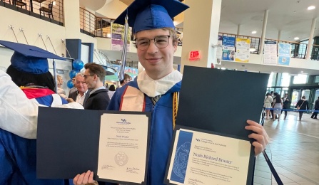 Noah Bruster holding up two degrees after commencement wearing a blue cap and gown. 