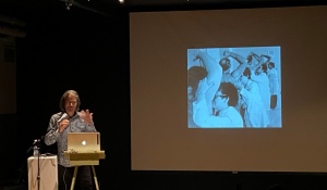 Photo of image projected on screen of a row people wearing scientist lab coats smelling subject arm pits with Scholars at Hallwalls presenter Paul Vanouse stnading at a lecturn speaking. 