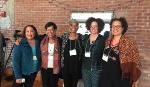 Photo of five women, arm-in-arm, wearing annual conference name tag lanyards in front of a brick wall from the Reclaiming Our Ancestors conference organized by the Humanities Institute. 