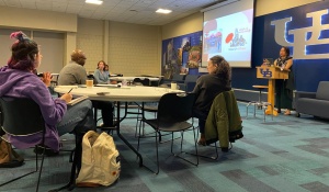 Photo of book talk attendees seated at round tables, with one attendee asking a question of the presenter Elena Shih with UB interlocking logo on wall in background. 