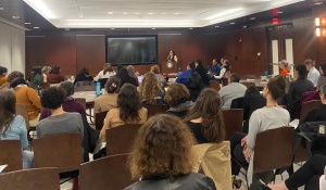 Photo of attendees seated in rows of chairs watching presenter. 