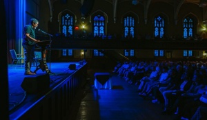 2024 Buffalo Humanities Festival spotlight speaker Percival Everett speaks on stage at a lectern with microphone under a spotlight, addressing a seated audience, cast in blue shadow, inside Asbury Hall, a converted church with stained glass windows. 