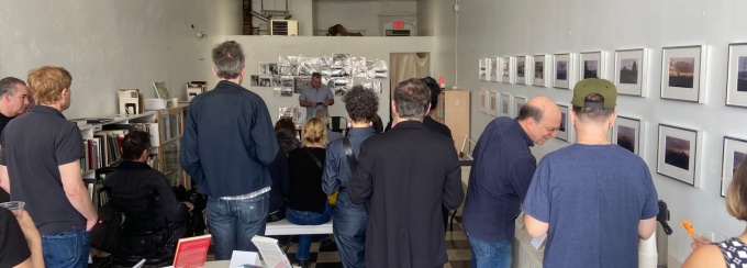 Photo of crowd in attendance at 2024 Spring Book Celebration, standing and listening to David Castillo provide introductions in a white-walled room with framed images on the wall, black-and-white checkered floor, with a round table with books on display stands. 