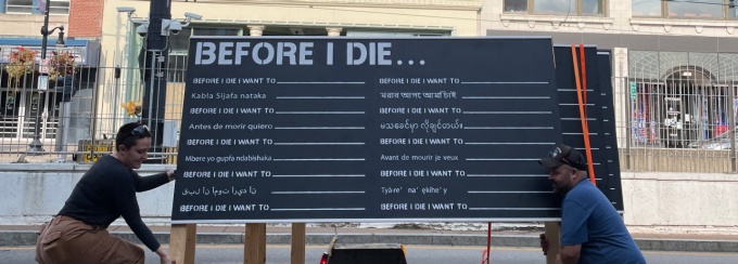 Photo of "Before I Die" interactive public art work chalk board on flat bed truck with two people lifting it for installation on Main Street in Buffalo, NY. 