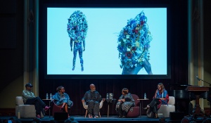 A panel discussion on a stage in a large theater setting, with a screen behind them displaying images of two figures covered in floral arrangements; 5 panelists sit in chairs and talk. The panelists pictured are Edreys Wajed, Victoria Udondian, Nick Cave, Celeste Lawson, and Claire Schneider. 