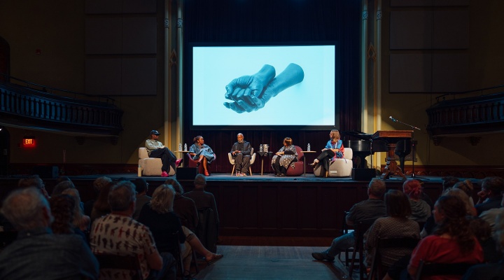 Photo of 5 people seated in arm chairs on a stage, with an image of a sculpture depicting two hands, one nestled into the palm of another projected on a screen above them. A seated crowd watches the interaction on stage. 