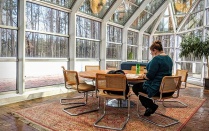 A person is seated at a round table with multiple chairs, reading papers on the table in an atrium-like glass walled and ceilinged space, surrounded by woods outside. 