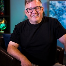 Marc Isaacs sitting on a chair in front of a blue background. 