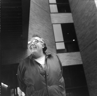 A man smiling and looking off to the side, standing outside of a building. American Studies professor John Mohawk, 1988; 18.5x18.3cm; Copyright 1988 University at Buffalo. 