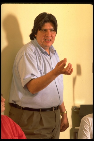 A man standing in front of a whiteboard in his classroom speaking to students and motioning with his hand. 