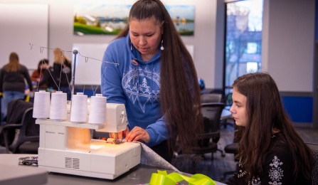 A woman standing behind a sewing machine demonstrating a sewing technique to a student who is sitting next to the sewing machine during a craft workshop. 