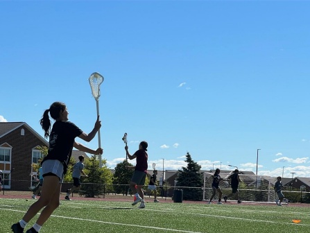 Students holding lacrosse sticks and running outside in a field during a lacrosse skills camp. 