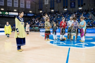 Zoom image: Community demonstration of cultural dance. Men are standing with hand rattles and women are standing away from them preparing to dance.