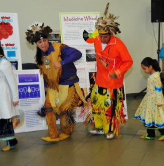 Zoom image: Two adult men standing and dancing during a community demonstration of cultural dance. A little girl is behind the men also dancing.