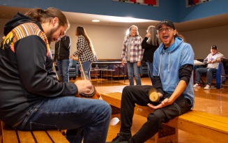 Zoom image: Two people sitting on wooden benches with hand rattles during a ssocial dance in the student union.