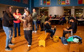 Zoom image: Group of people standing and holding hands while completing a "Rabbit Dance." Two individuals are sitting on wooden benches as well.