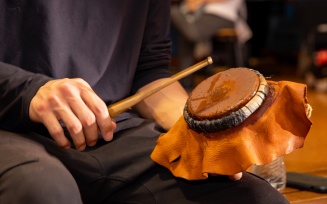 Zoom image: Individual sitting with a water drum, which is being used in a Haudenosaunee Social Dance. One hand holds the drum and the other holds a stick.
