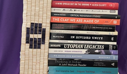 Staff of books. To the left of the books, is a wampum belt with a UB sign on top of it, followed by a purple Haudenosaunee flag at the top. 