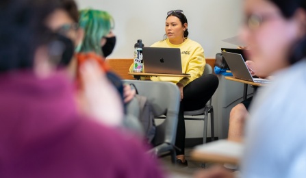 Students sitting at desks in a classroom. The students are the front of the image are blurred and the camera is focused on a student in a yellow sweatshirt with a laptop and water bottle in front of her. 