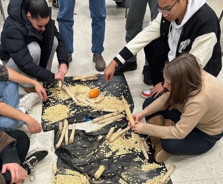 A black tarp on the floor covered with pieces of corns. Hands from students can be seen reaching for the pieces of corn.