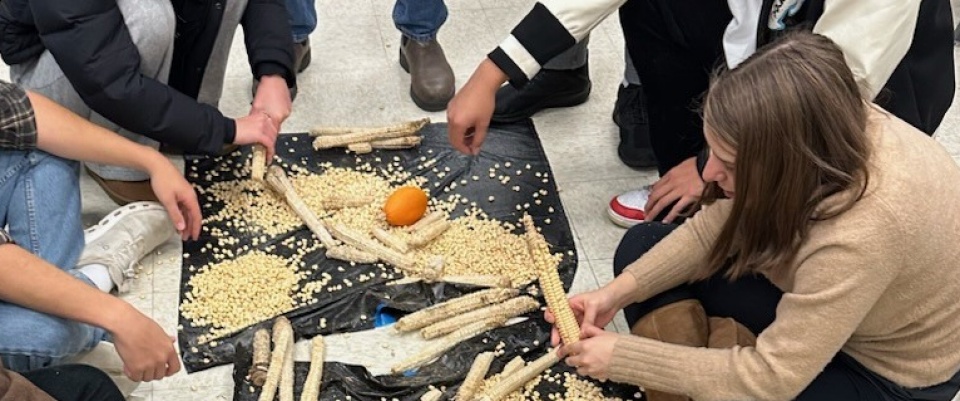 A black tarp on the floor covered with pieces of corns. Hands from students can be seen reaching for the pieces of corn. 