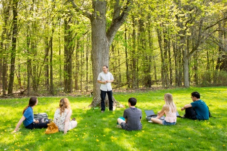 Jason Corwin lecturing in his land-based learning class. Students are sitting in a circle outside on the grass, while a man stands in front of a tree talking to them. 