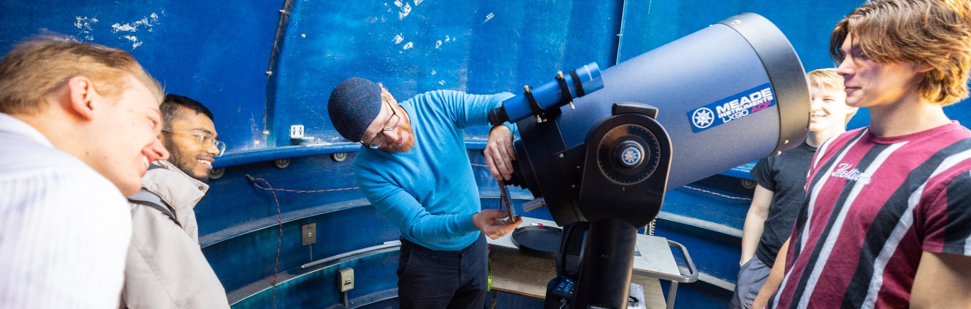 Instructor adjusts a large telescope while several students stand around inside a blue observatory dome on the roof of Fronczak Hall. 