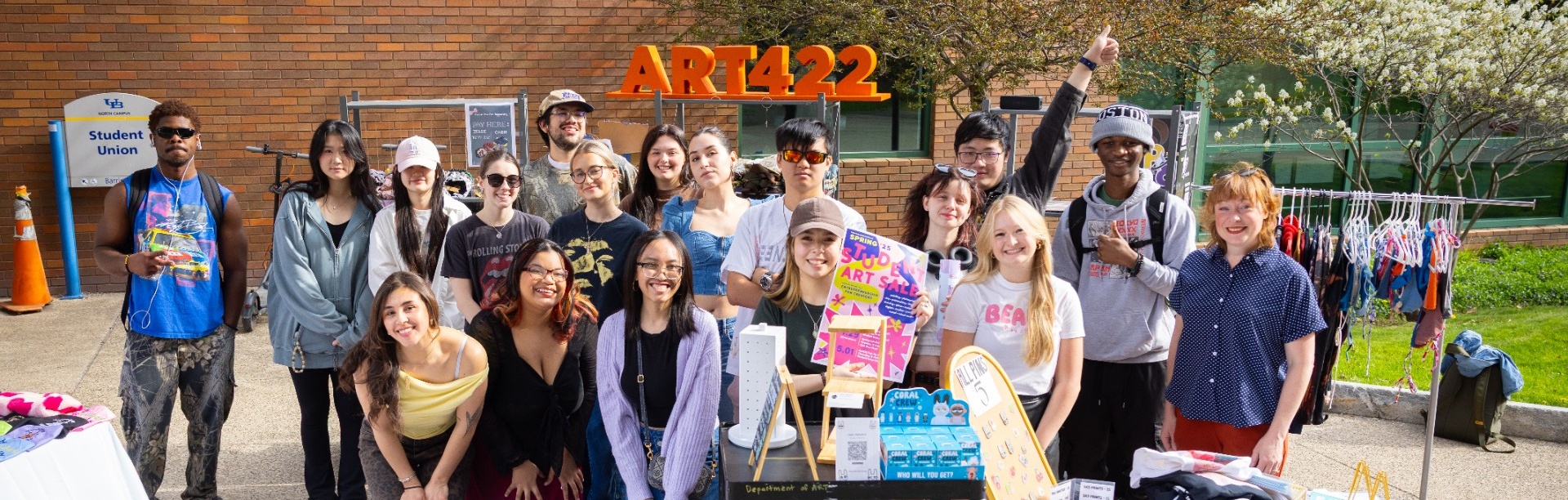 Group of students from an ART 422 class stand behind outdoor pop-up tables displaying handmade artwork and products. Signs include “ART422” in large orange letters behind them and a banner reading “PEACHYDEADLACK HANDMADE DESIGNS.” A poster on one table reads “Student Art Sale.” Tables are filled with prints, crafts, and small items for sale as students present their work on campus.