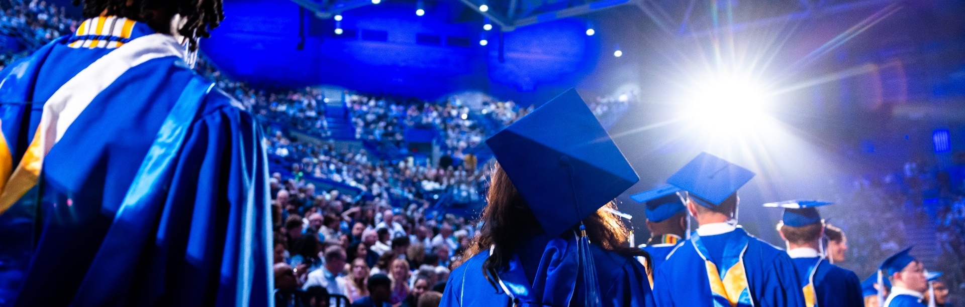 The image is filled with rows of people wearing bright blue graduation caps (mortarboards) and matching gowns. The caps form a repeating geometric pattern across the entire scene. Many caps are plain blue, while others are decorated with colorful designs, messages, or embellishments, adding variation throughout the crowd. 