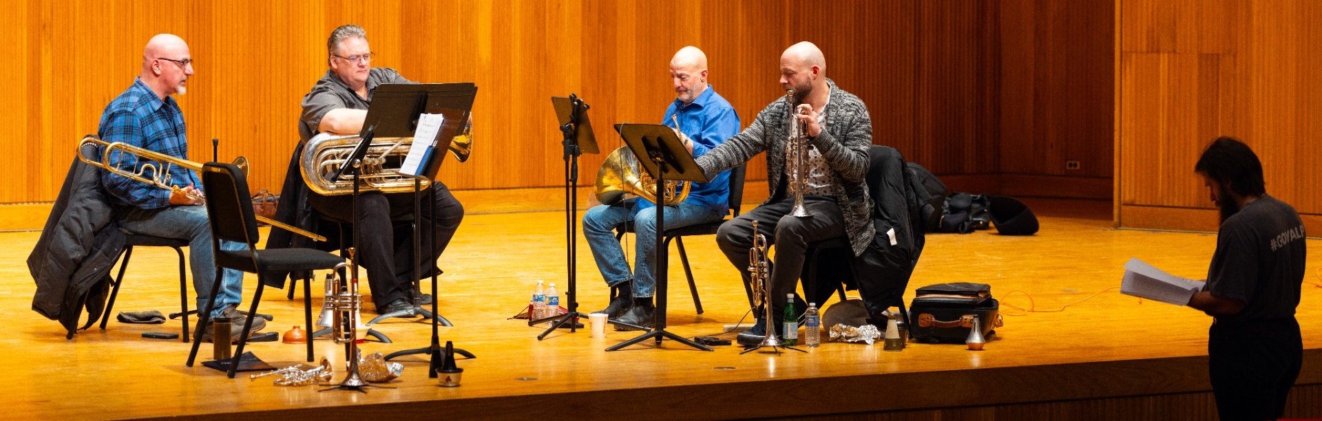 A brass ensemble rehearses on a concert hall stage while a student composer stands nearby reviewing a score. The scene captures a collaborative music workshop in which professional musicians perform student-composed works and provide artistic feedback. 