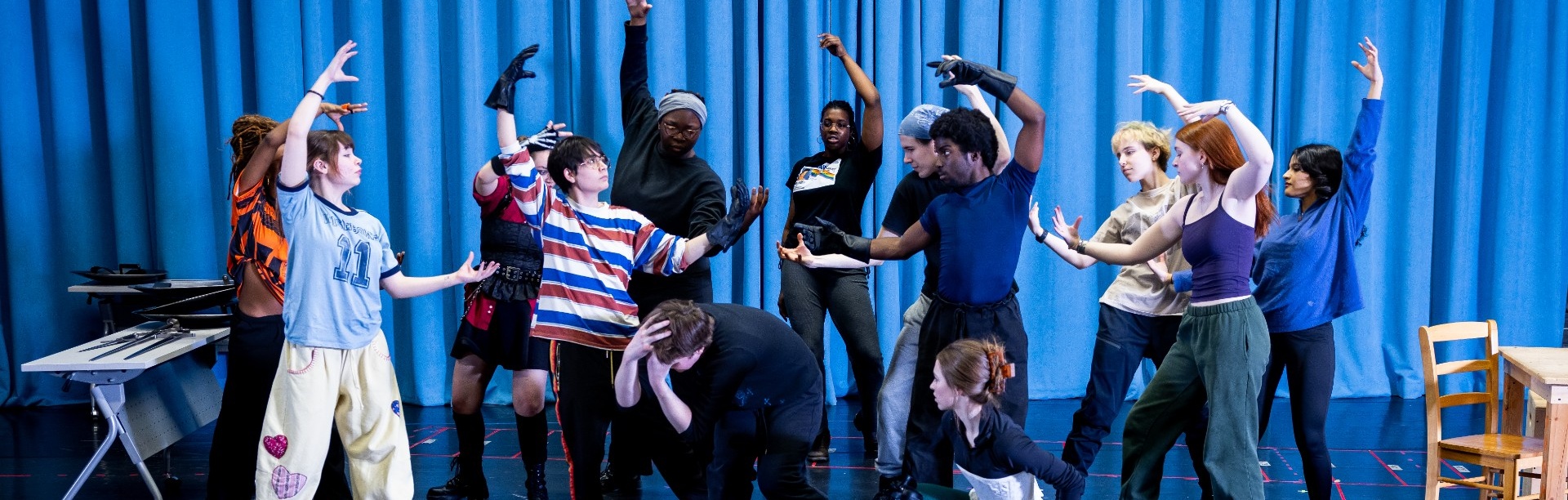 Theater students rehearse a scene on a black box stage, using expressive poses and movement while surrounded by rehearsal props, stage markings, and a blue curtain backdrop. 