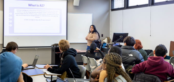 A college classroom is shown during a class lecture on artificial intelligence. Students sit at individual desks facing the front of the room, where a student presenter stands beside a projected slide. At the front of the classroom, a professor stands near a lectern and computer workstation, addressing the class. Behind her, a large projection screen displays a slide titled: &ldquo;What is AI?&rdquo;. 