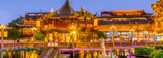 Illuminated traditional Chinese-style buildings and bridges reflected in a pond at dusk. 