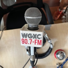 A close-up of a WGX C 90.7 FM studio microphone on a desk, with headphones resting nearby. 