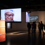 Installation view of Benson's exhibit showing an orange oil barrel in foreground, three videos projected onto hanging screens arranged throughout the space, and several silhouetted visitors. 