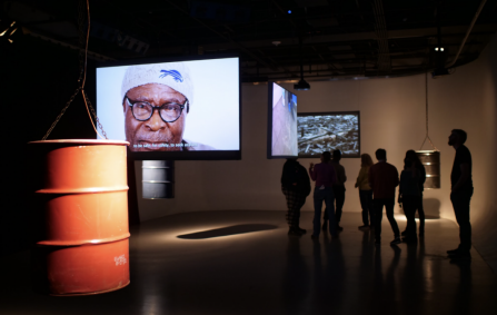 Installation view of Benson's exhibit showing an orange oil barrel in foreground, three videos projected onto hanging screens arranged throughout the space, and several silhouetted visitors. 